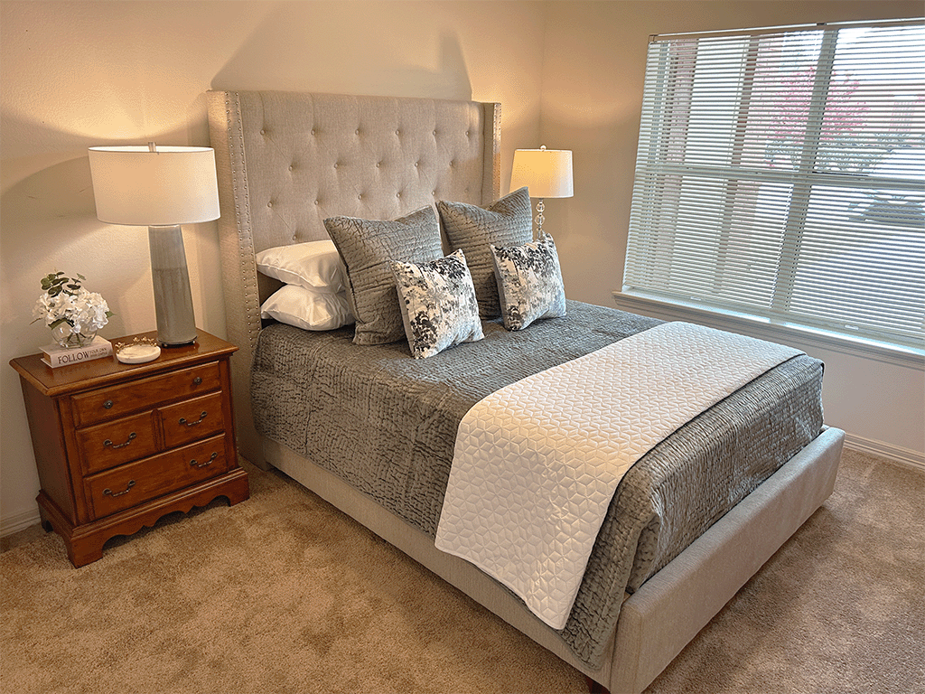 A bedroom with a large bed, a nightstand with a lamp, and a window with blinds.